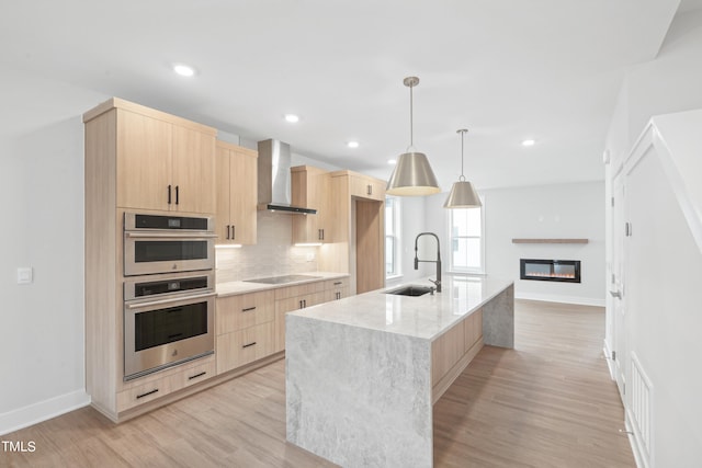 kitchen with wall chimney range hood, sink, a center island with sink, decorative light fixtures, and light brown cabinets