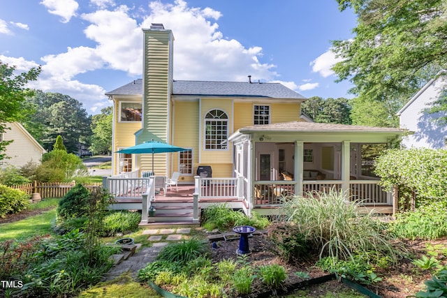 rear view of property featuring a sunroom, a chimney, a wooden deck, fence, and roof with shingles