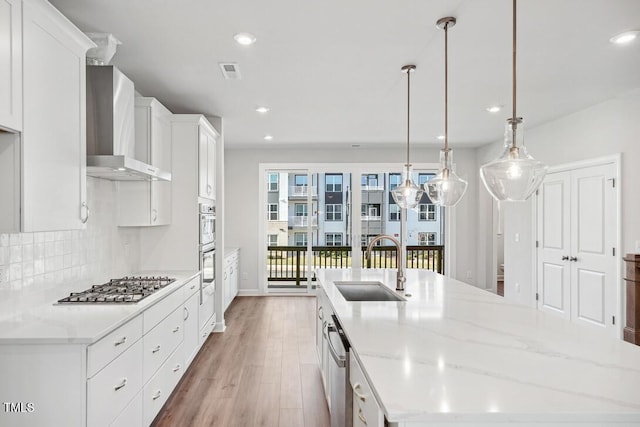 kitchen featuring white cabinetry, stainless steel appliances, wall chimney range hood, pendant lighting, and a center island with sink