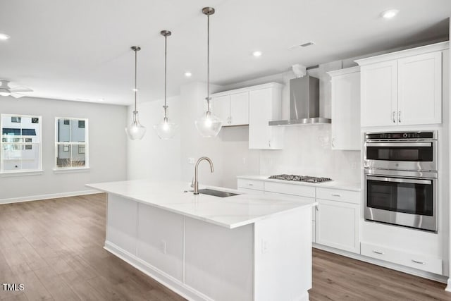 kitchen featuring sink, wall chimney exhaust hood, an island with sink, decorative light fixtures, and stainless steel appliances