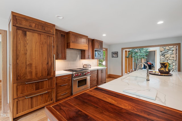 kitchen with sink, backsplash, high end stainless steel range oven, light stone counters, and light hardwood / wood-style flooring