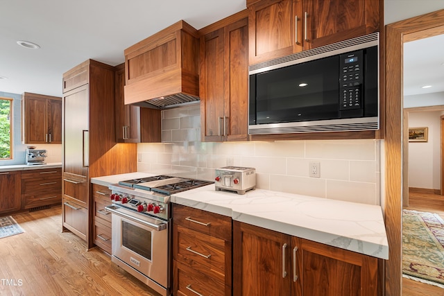 kitchen featuring appliances with stainless steel finishes, backsplash, light stone countertops, custom range hood, and light wood-type flooring