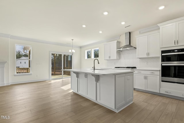 kitchen with stainless steel double oven, white cabinetry, an island with sink, and wall chimney exhaust hood