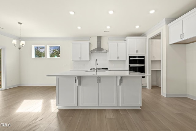 kitchen featuring an island with sink, white cabinets, black double oven, wall chimney range hood, and backsplash