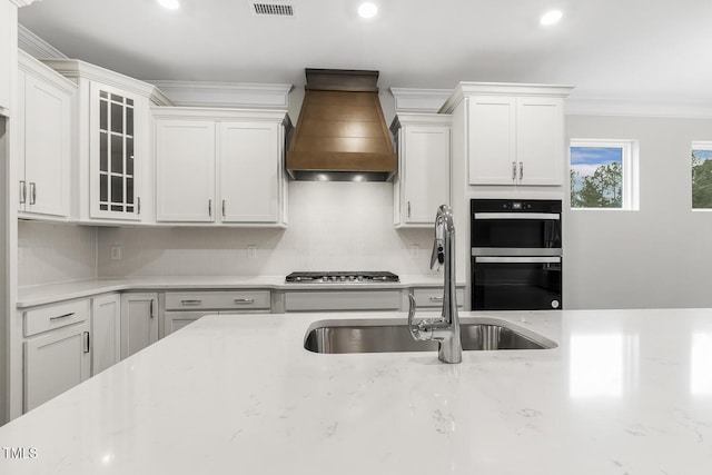 kitchen with sink, white cabinets, custom exhaust hood, black double oven, and crown molding