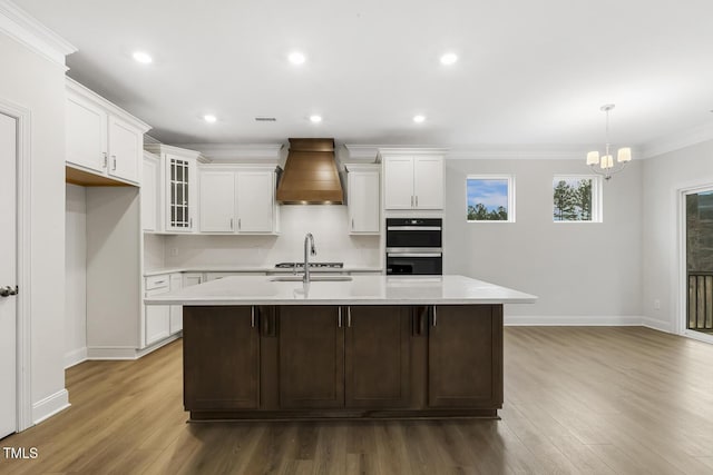 kitchen featuring custom exhaust hood, white cabinetry, a kitchen island with sink, and sink