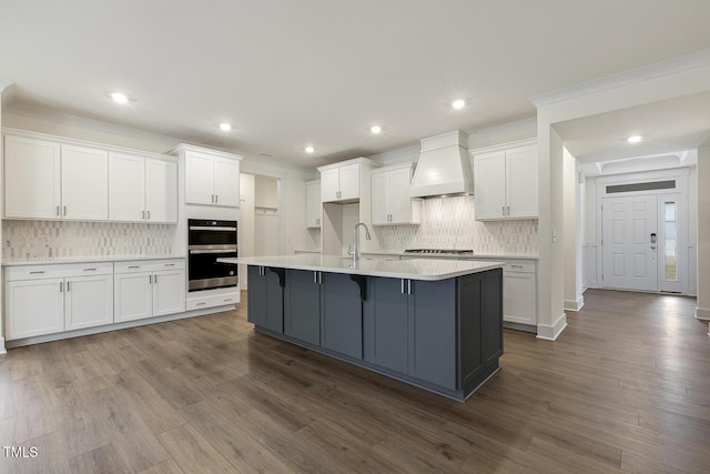 kitchen with sink, custom exhaust hood, wood-type flooring, an island with sink, and white cabinets