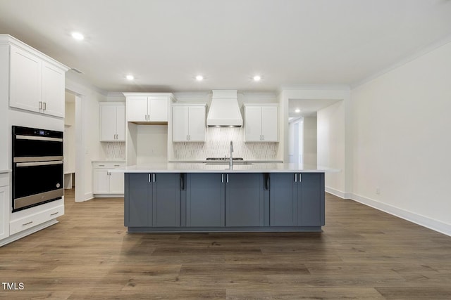 kitchen featuring white cabinetry, double oven, a kitchen island with sink, and custom range hood