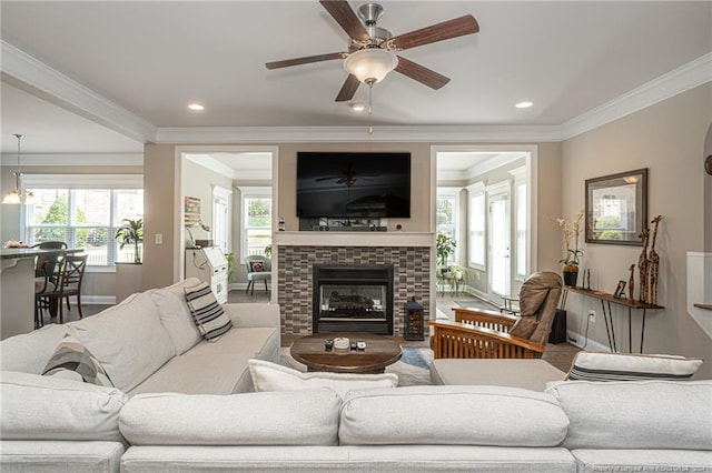 living room with ceiling fan with notable chandelier, crown molding, and a stone fireplace