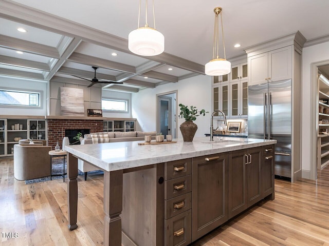 kitchen featuring stainless steel built in fridge, a center island with sink, light stone countertops, light wood-type flooring, and decorative light fixtures