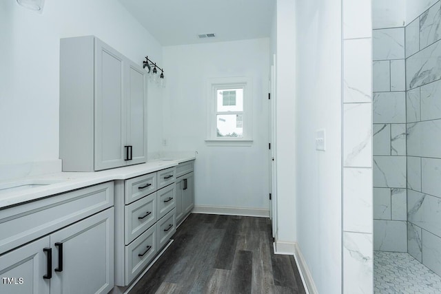 bathroom featuring wood finished floors, visible vents, baseboards, a tile shower, and double vanity