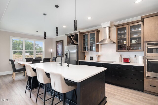 kitchen with a kitchen island with sink, wall chimney range hood, hanging light fixtures, light hardwood / wood-style floors, and stainless steel appliances