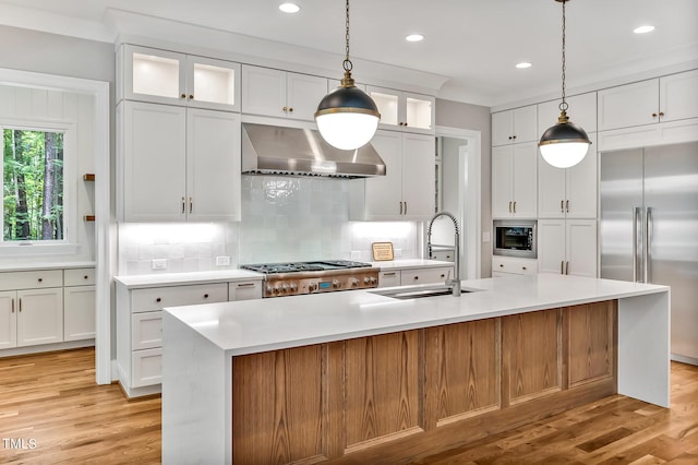 kitchen with sink, built in appliances, wall chimney exhaust hood, decorative backsplash, and an island with sink