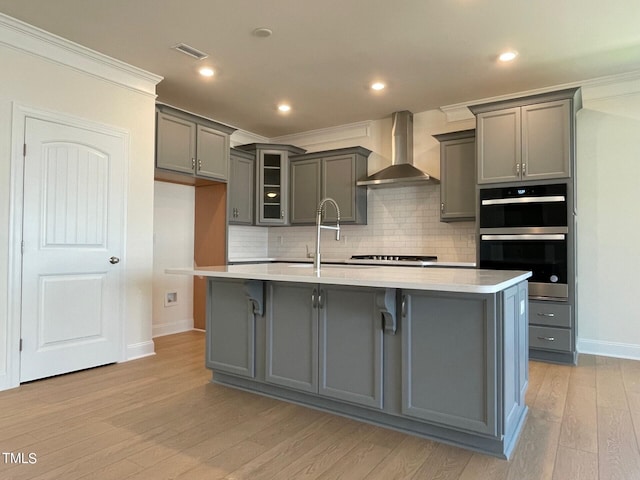 kitchen featuring wall chimney range hood, gray cabinets, a center island with sink, crown molding, and double oven