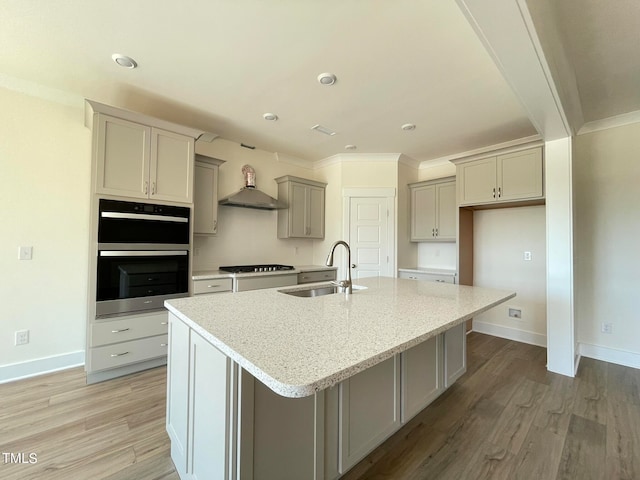 kitchen featuring a center island with sink, sink, multiple ovens, wall chimney exhaust hood, and light stone counters