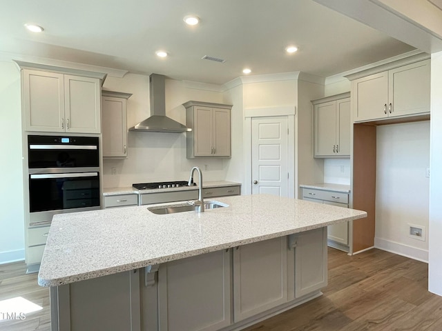 kitchen with light stone countertops, multiple ovens, a center island with sink, and wall chimney exhaust hood