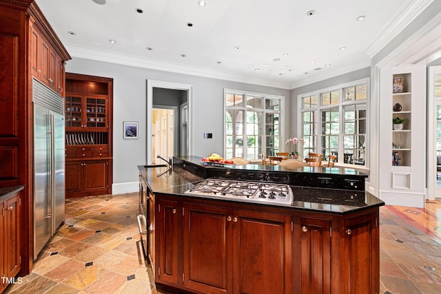 kitchen with built in shelves, dark stone counters, a kitchen island with sink, appliances with stainless steel finishes, and ornamental molding