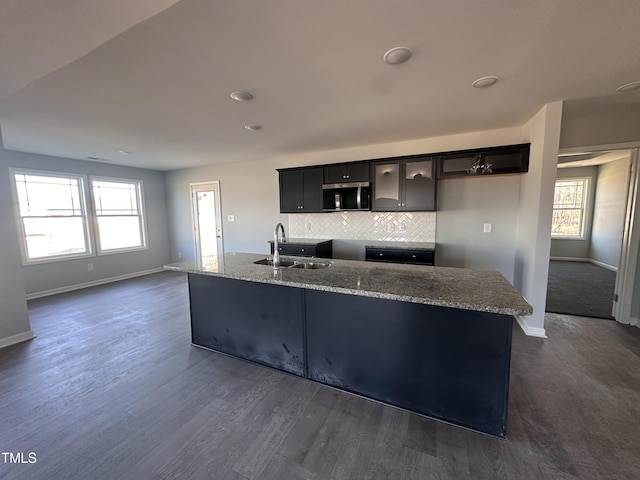 kitchen with backsplash, sink, light stone countertops, an island with sink, and dark wood-type flooring