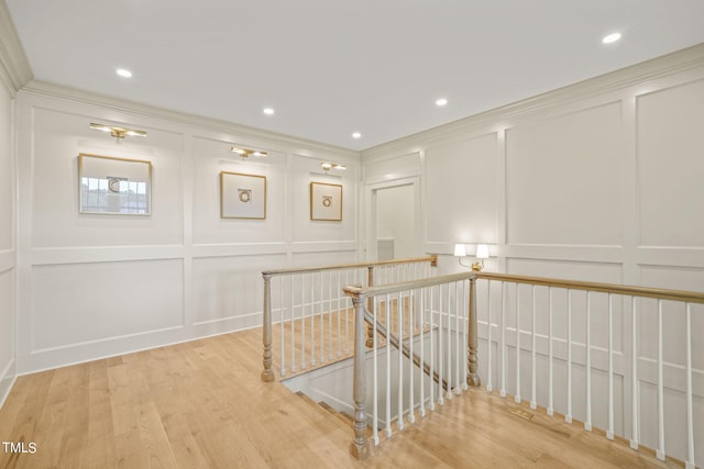 hallway featuring light wood finished floors, crown molding, a decorative wall, and an upstairs landing