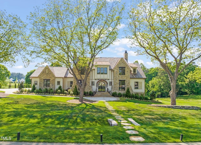 english style home featuring stone siding, a chimney, and a front yard
