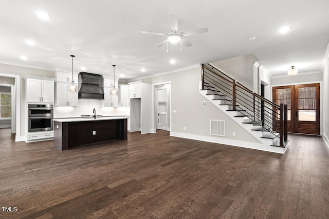 kitchen with pendant lighting, double oven, white cabinetry, custom range hood, and a center island with sink