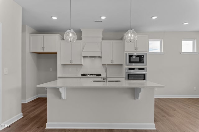 kitchen featuring white cabinetry, stainless steel oven, an island with sink, and hanging light fixtures