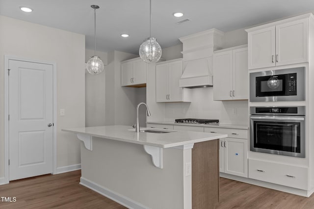 kitchen with custom exhaust hood, stainless steel oven, sink, white cabinetry, and an island with sink