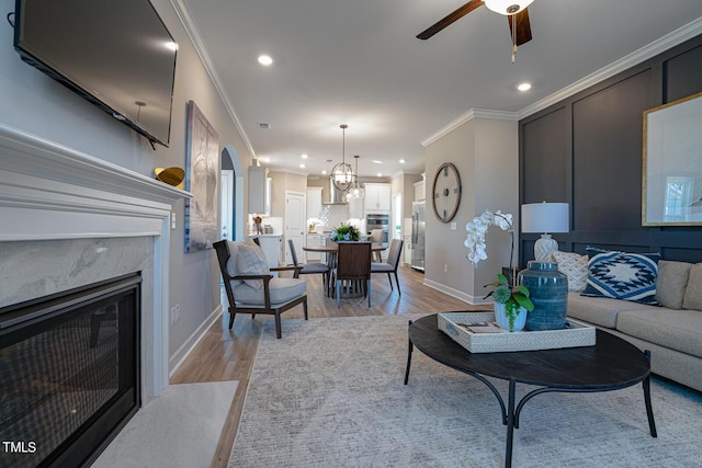 living room with light wood-style flooring, ceiling fan with notable chandelier, crown molding, and a high end fireplace