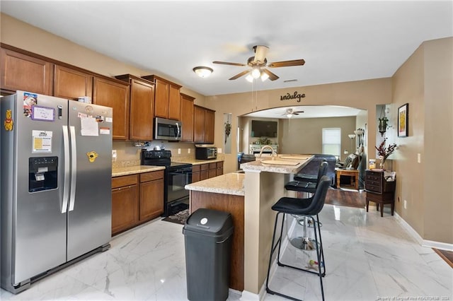 kitchen featuring a breakfast bar, a kitchen island with sink, ceiling fan, light stone countertops, and appliances with stainless steel finishes