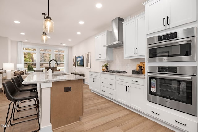 kitchen with wall chimney range hood, sink, a kitchen island with sink, white cabinets, and light stone counters