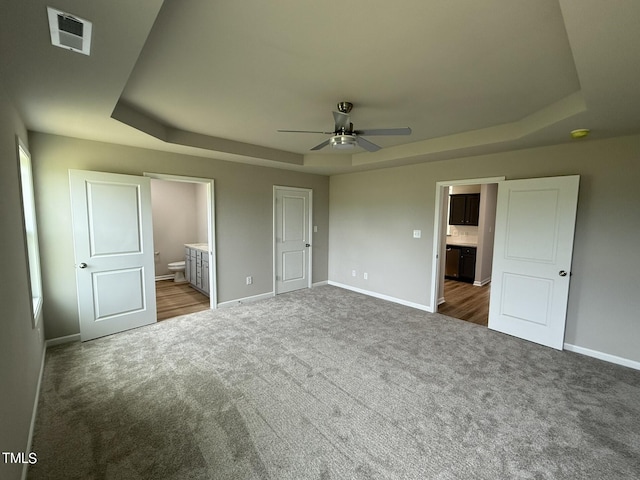 unfurnished bedroom with dark carpet, ensuite bath, ceiling fan, and a raised ceiling