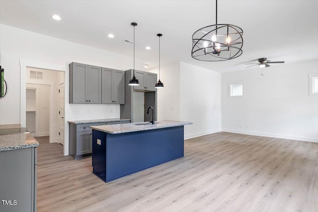 kitchen featuring sink, light stone counters, decorative light fixtures, a center island with sink, and gray cabinets