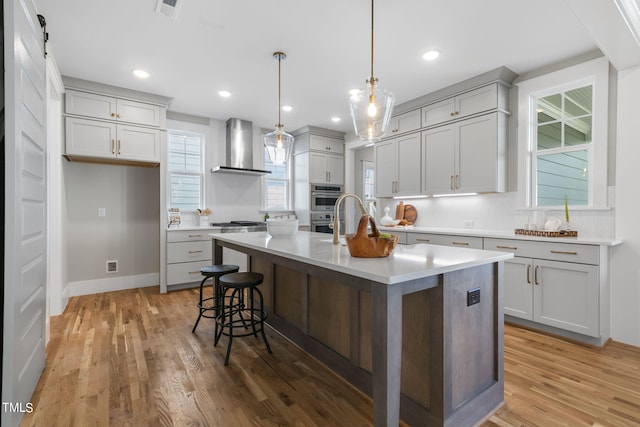 kitchen featuring appliances with stainless steel finishes, wood-type flooring, wall chimney exhaust hood, decorative light fixtures, and a kitchen island with sink