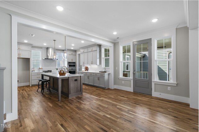 kitchen featuring a center island with sink, pendant lighting, a kitchen breakfast bar, gray cabinetry, and wall chimney exhaust hood