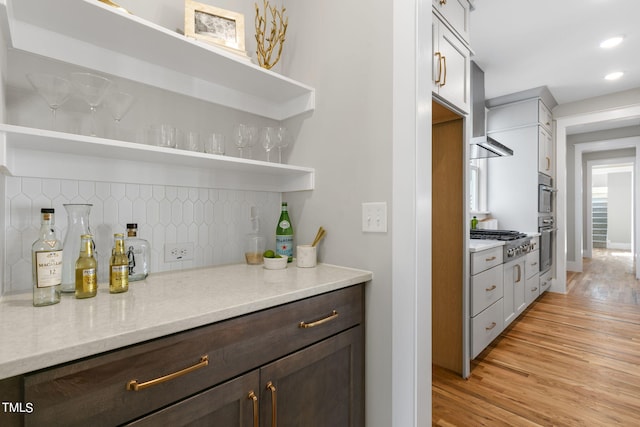 bar with appliances with stainless steel finishes, light stone countertops, light hardwood / wood-style floors, dark brown cabinetry, and decorative backsplash