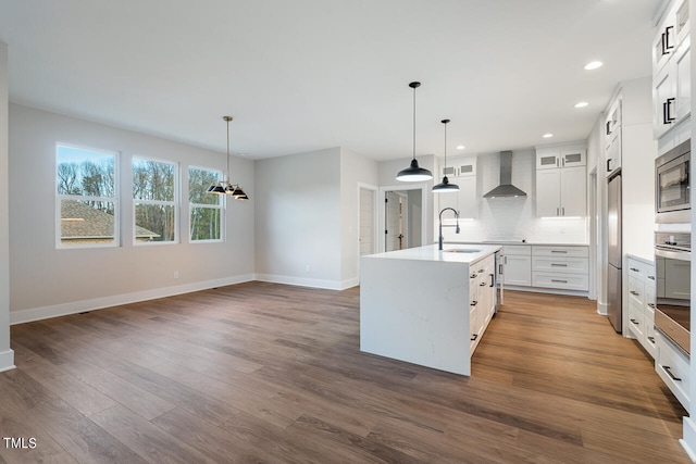 kitchen with hardwood / wood-style flooring, wall chimney range hood, white cabinets, appliances with stainless steel finishes, and a kitchen island with sink