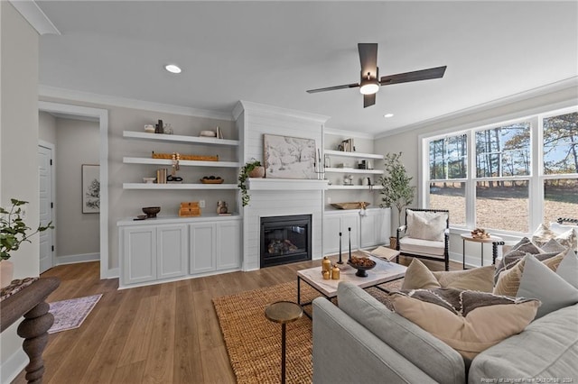 living room with built in shelves, ceiling fan, a large fireplace, crown molding, and light hardwood / wood-style floors