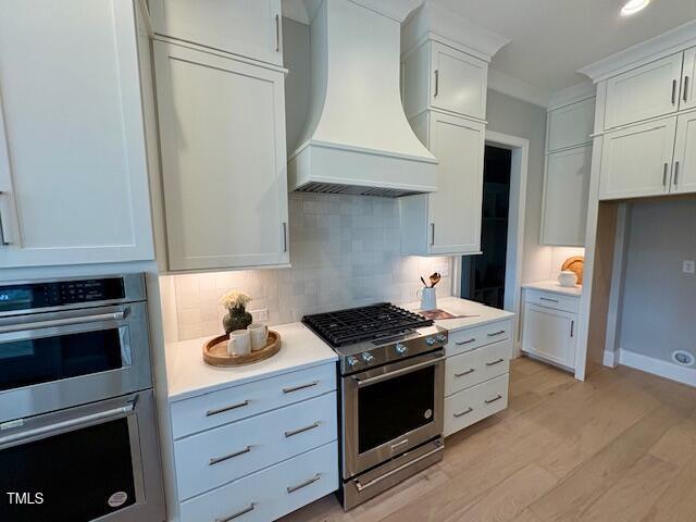 kitchen featuring light countertops, decorative backsplash, appliances with stainless steel finishes, light wood-type flooring, and premium range hood