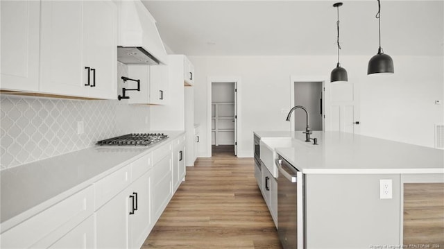 kitchen featuring appliances with stainless steel finishes, light wood-type flooring, premium range hood, decorative light fixtures, and a center island with sink