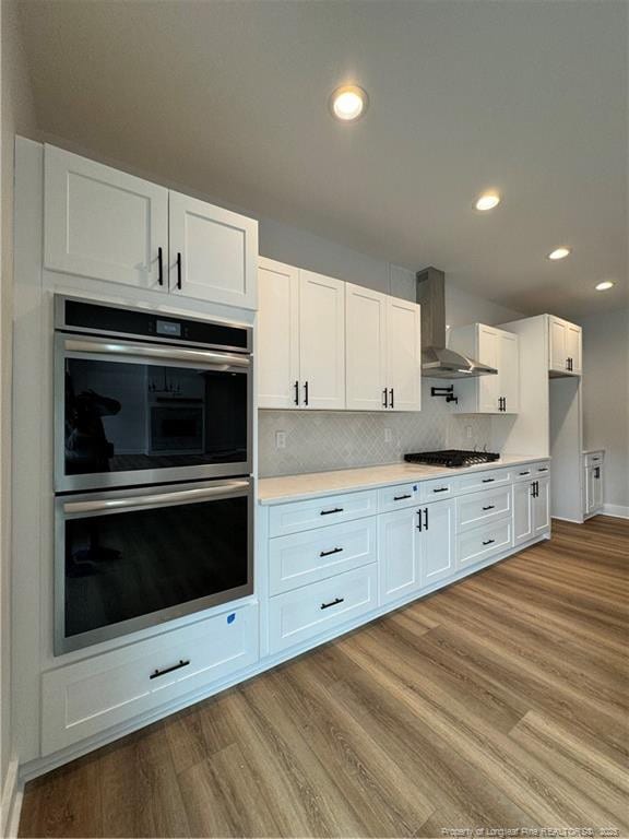 kitchen with white cabinets, wall chimney exhaust hood, light wood-style flooring, appliances with stainless steel finishes, and light countertops