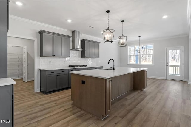 kitchen featuring wall chimney exhaust hood, stainless steel gas cooktop, sink, gray cabinetry, and a center island with sink