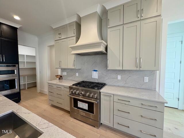 kitchen with appliances with stainless steel finishes, light stone counters, tasteful backsplash, custom range hood, and light wood-type flooring