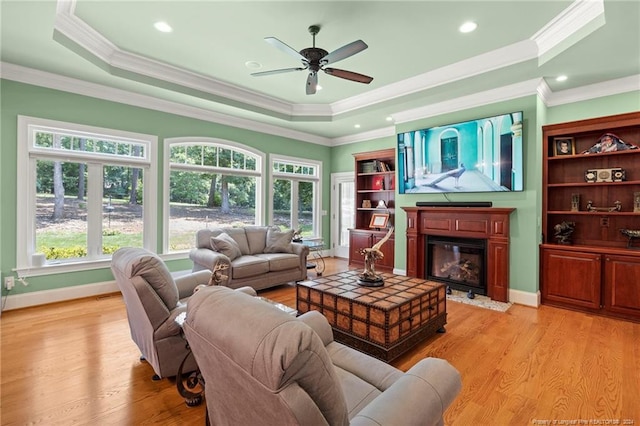 living area featuring light wood finished floors, ornamental molding, ceiling fan, a glass covered fireplace, and a raised ceiling