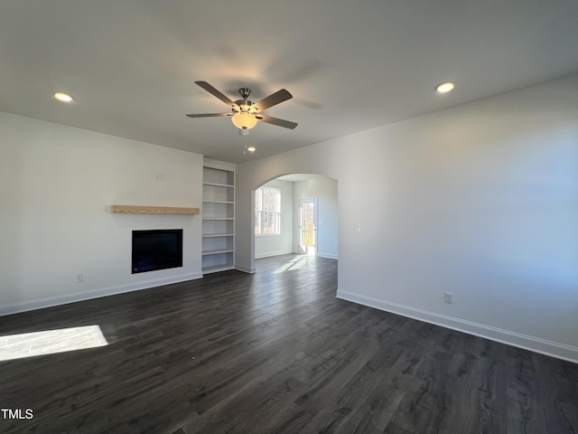 unfurnished living room featuring built in shelves, ceiling fan, and dark wood-type flooring