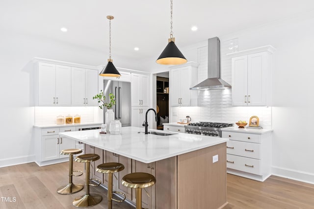 kitchen featuring sink, high end fridge, white cabinetry, a center island with sink, and wall chimney exhaust hood