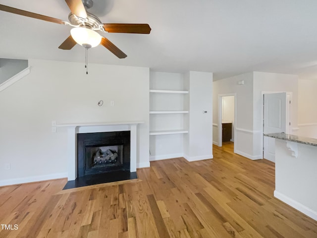 unfurnished living room featuring built in shelves, ceiling fan, and light wood-type flooring