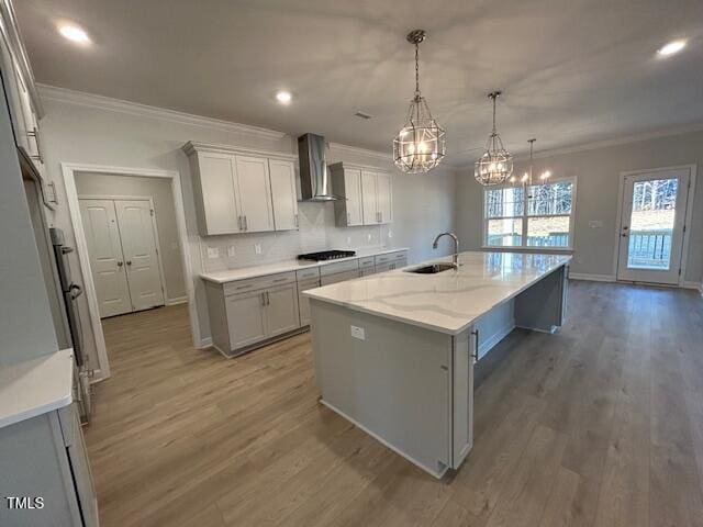 kitchen with gas cooktop, wall chimney exhaust hood, a kitchen island with sink, sink, and pendant lighting