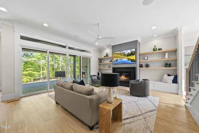 living area featuring light wood-type flooring, a glass covered fireplace, stairway, ceiling fan, and ornamental molding