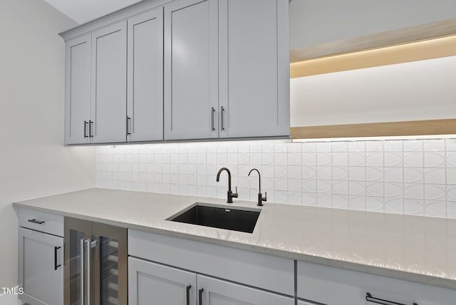 kitchen with wine cooler, a sink, backsplash, light stone counters, and gray cabinets