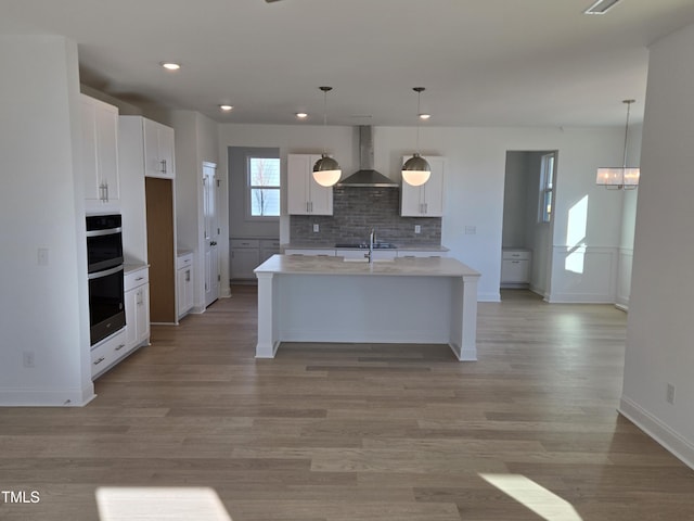 kitchen featuring white cabinetry, wall chimney range hood, light hardwood / wood-style flooring, an island with sink, and pendant lighting